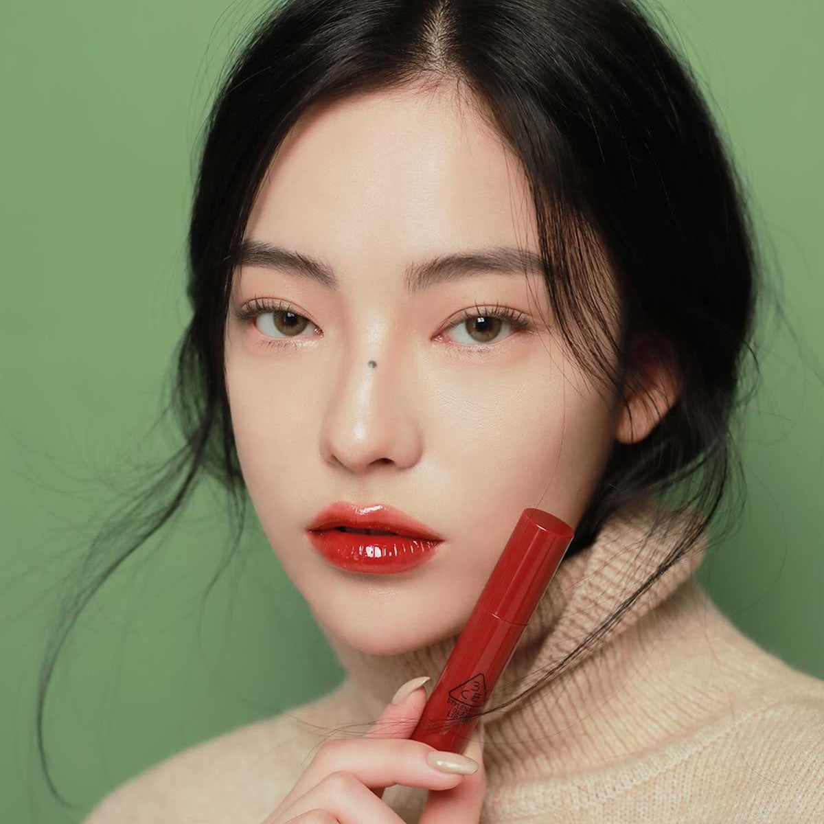 3CE Glaze Lip Tint 7 Shades, femme applique un rouge à lèvres brillant sur fond vert, look maquillage 3CE