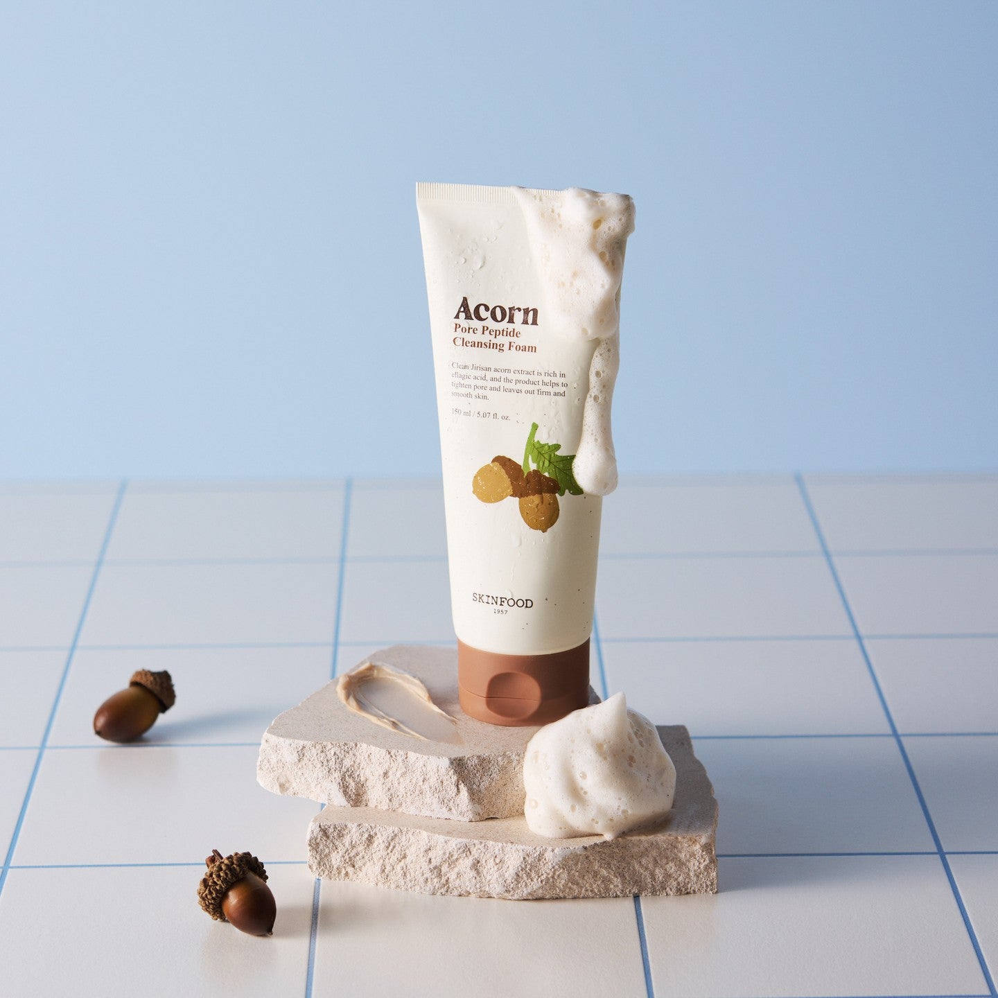 SKINFOOD Acorn Pore Peptide Cleansing Foam 150ml sur fond bleu avec mousse, éclats de pierre et noisettes