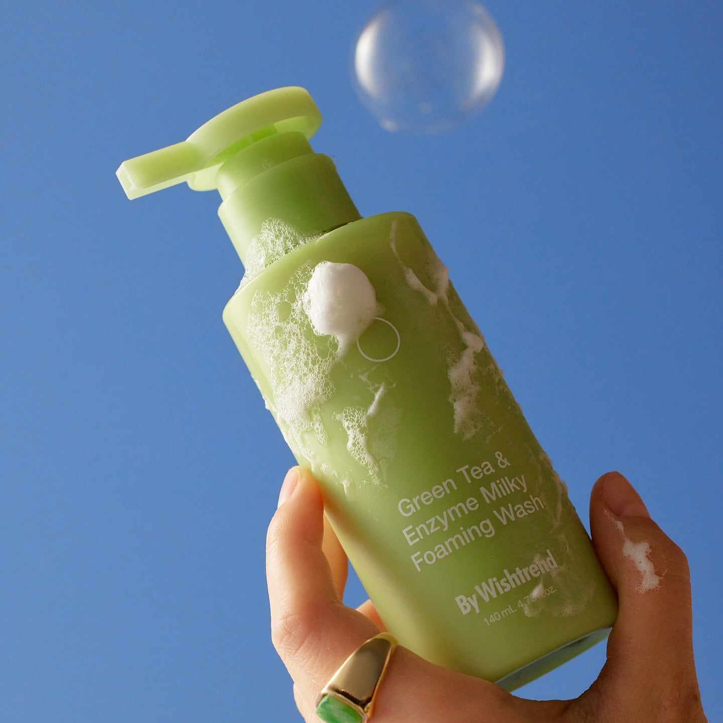 BY WISHTREND Green Tea & Enzyme Milky Foaming Wash 140ml : flacon vert en main, mousse sur fond bleu.