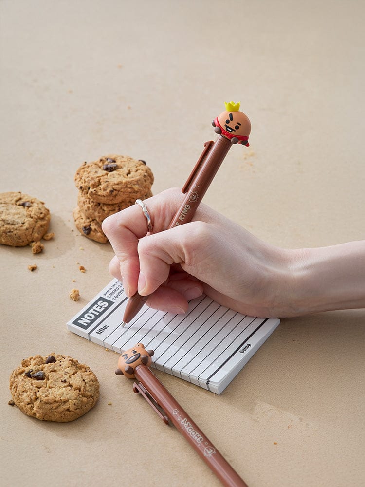 BT21 SHOOKY THE KING & BAKY FIGURE PEN LINE FRIENDS : stylo figurine sur carnet, main tenant le stylo, biscuits en arrière-plan.