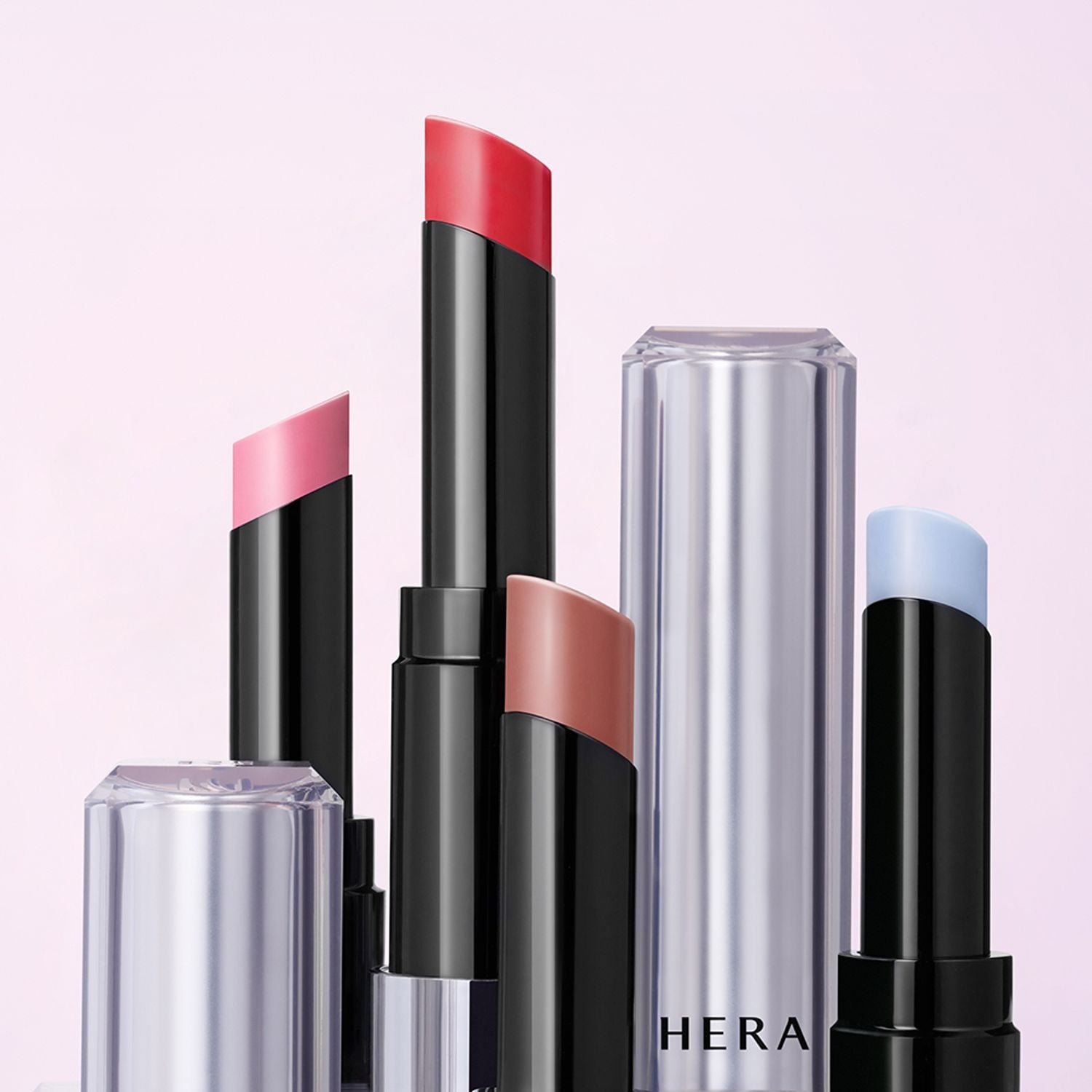 Décris l'image en français: HERA Sensual Nude Balm 3.5g, rouge à lèvres nude HERA sur fond rose, pack noir et tube transparent.