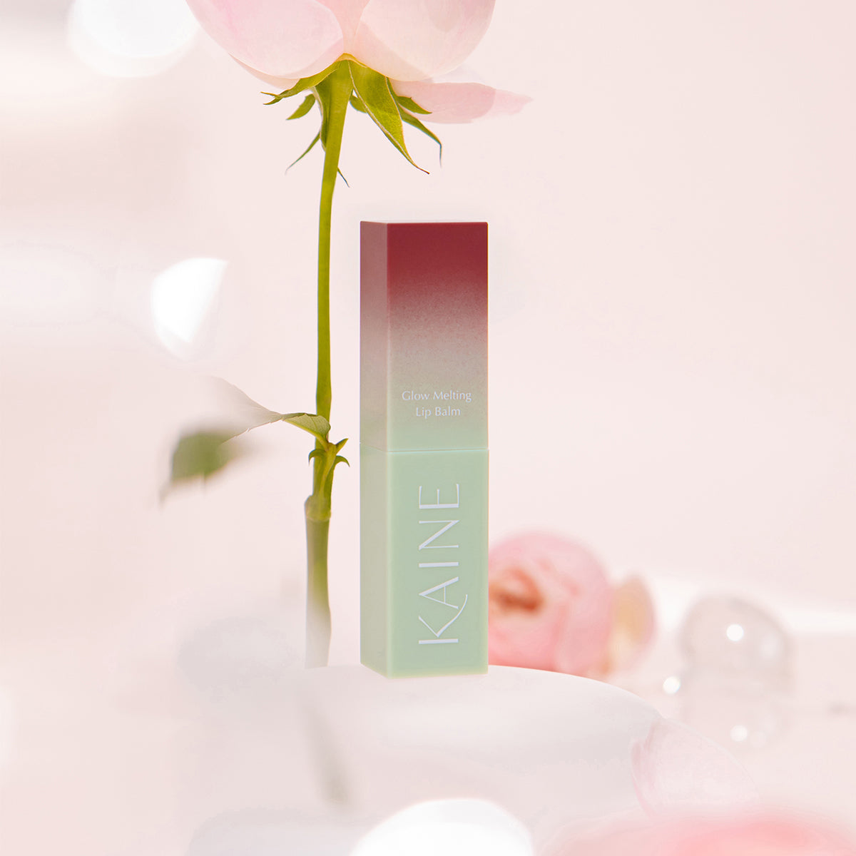 KAINE Glow Melting Lip Balm de la marque KAINE, flacon vert posé sur pétales roses, fond lumineux, esthétique douce.