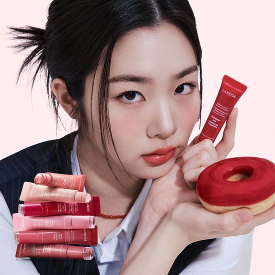 Décris l'image en français : LANEIGE Glaze Craze Tinted Lip Serum, femme tient le tube rouge et un donut, fond rose.