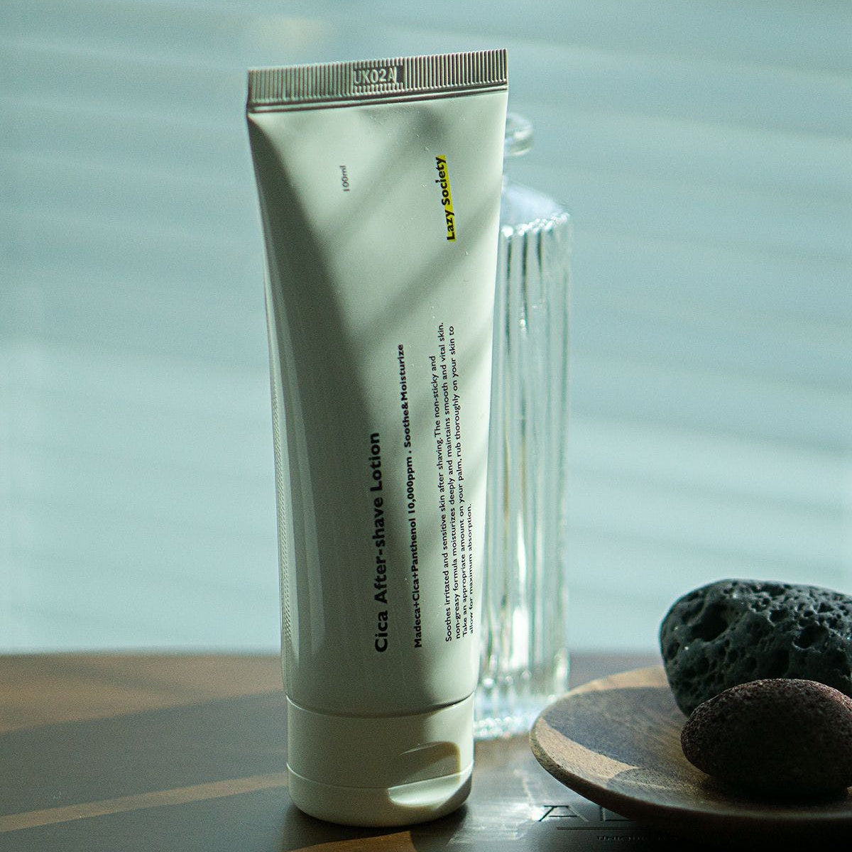 LAZY SOCIETY Cica Aftershave Lotion 100ml en tube vert sur fond spa, pierres et serviette, crème apaisante.