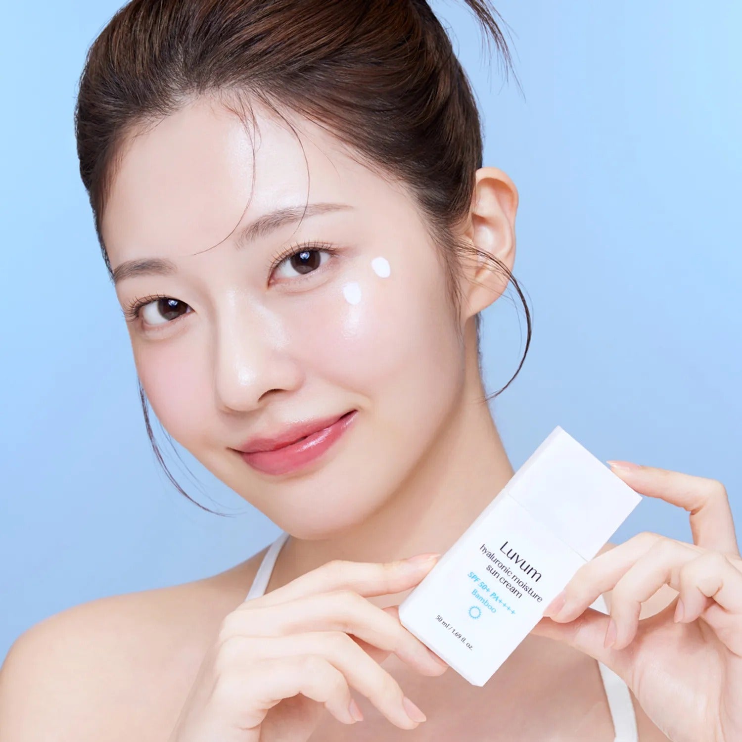Décris l'image en français: LUVUM Bamboo Hyaluronic Acid Moisture Sunscreen 30ml, femme au selfie, tube à la main, fond bleu.