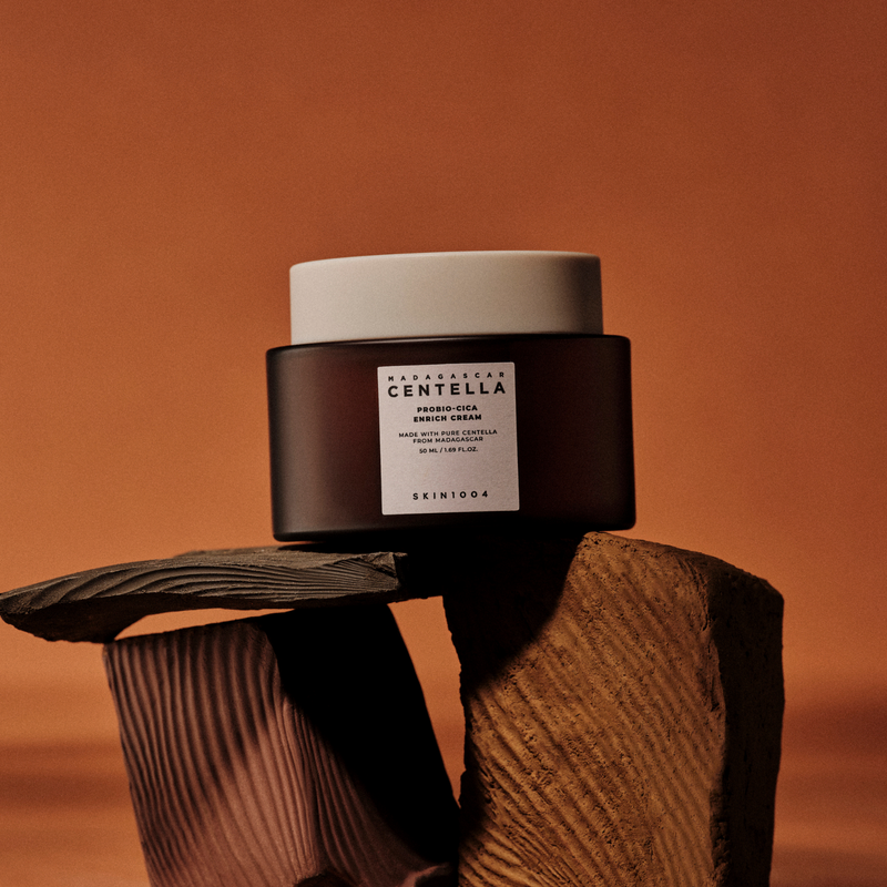SKIN1004 Madagascar Centella Probio-cica Enrich Cream 50ml, pot cosmétique sur tronc d’arbre, fond orange chaud.