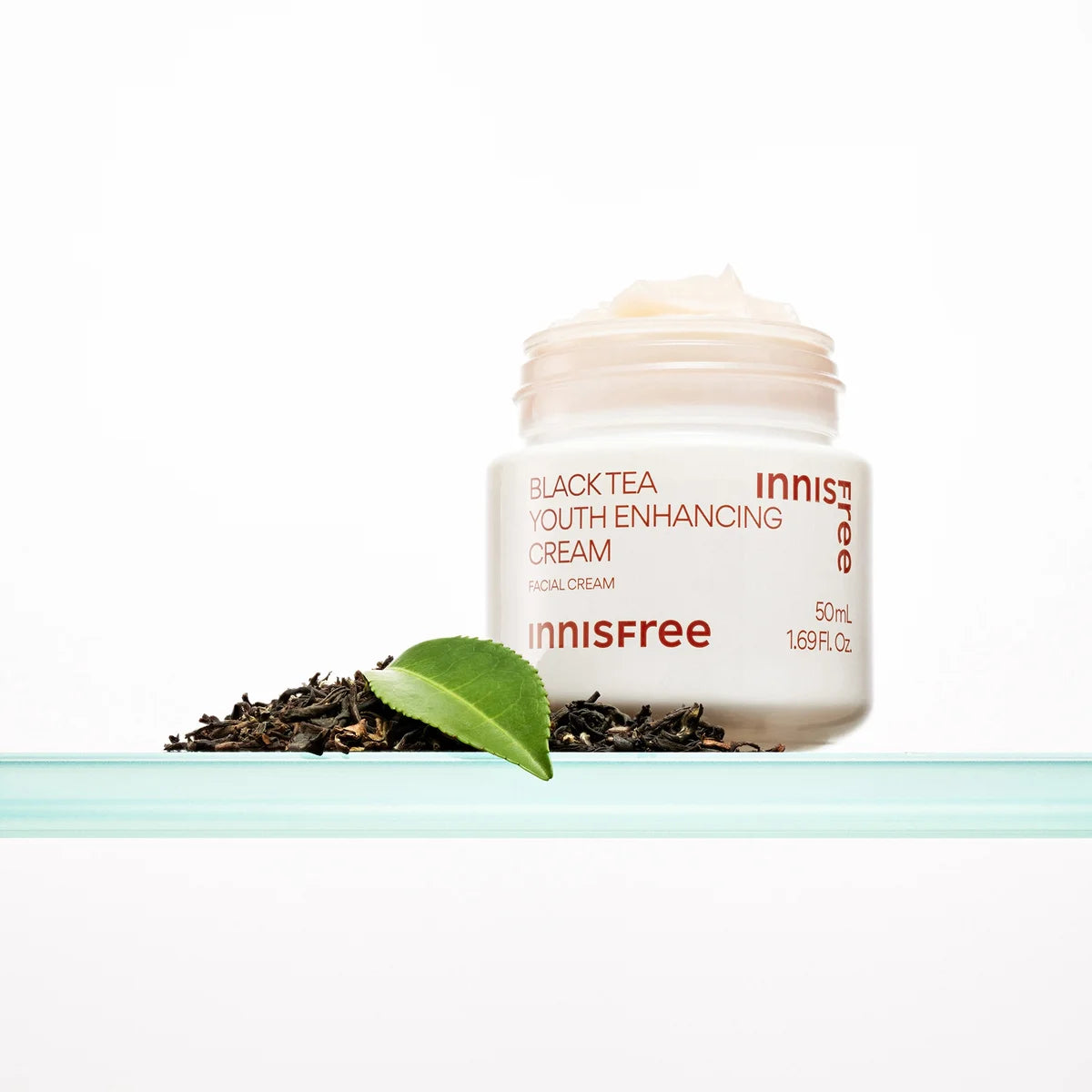 Décris l'image en français : INNISFREE Black Tea Youth Enhancing Cream 50ml, pot de crème sur fond blanc avec feuilles de thé noir.