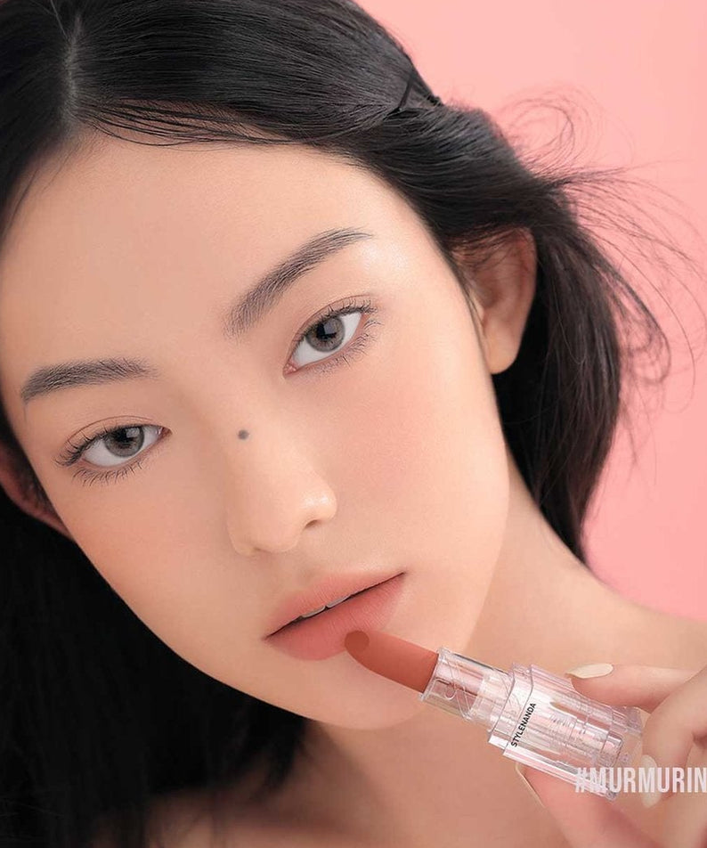 3CE Soft Matte Lipstick de la marque 3CE, modèle maquillée tenant le rouge à lèvres sur fond rose.