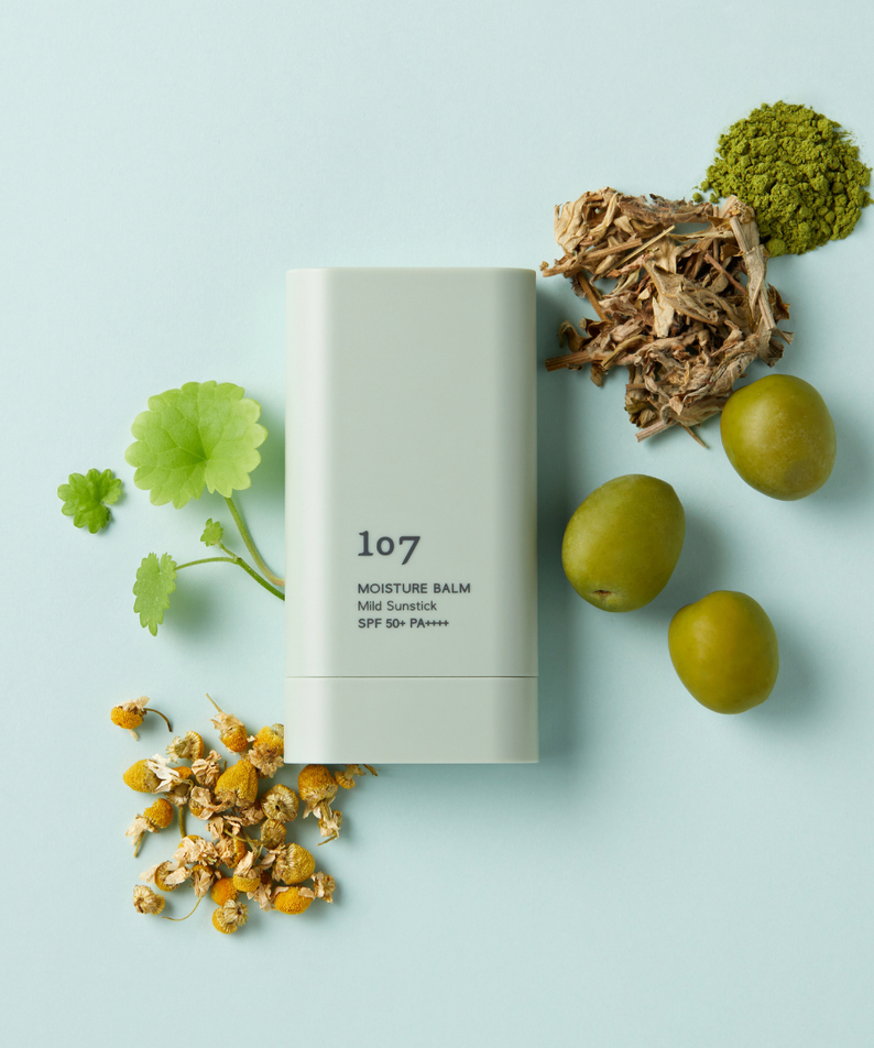 107 Moisture Balm Mild Sun Stick SPF50+PA++++ avec ingrédients naturels sur fond pastel vert.