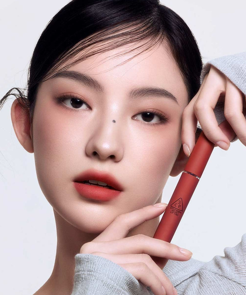 Gros plan sur 3CE Velvet Lip Tint de la marque 3CE, lèvres et tube rouge, maquillage et mains sur fond clair.
