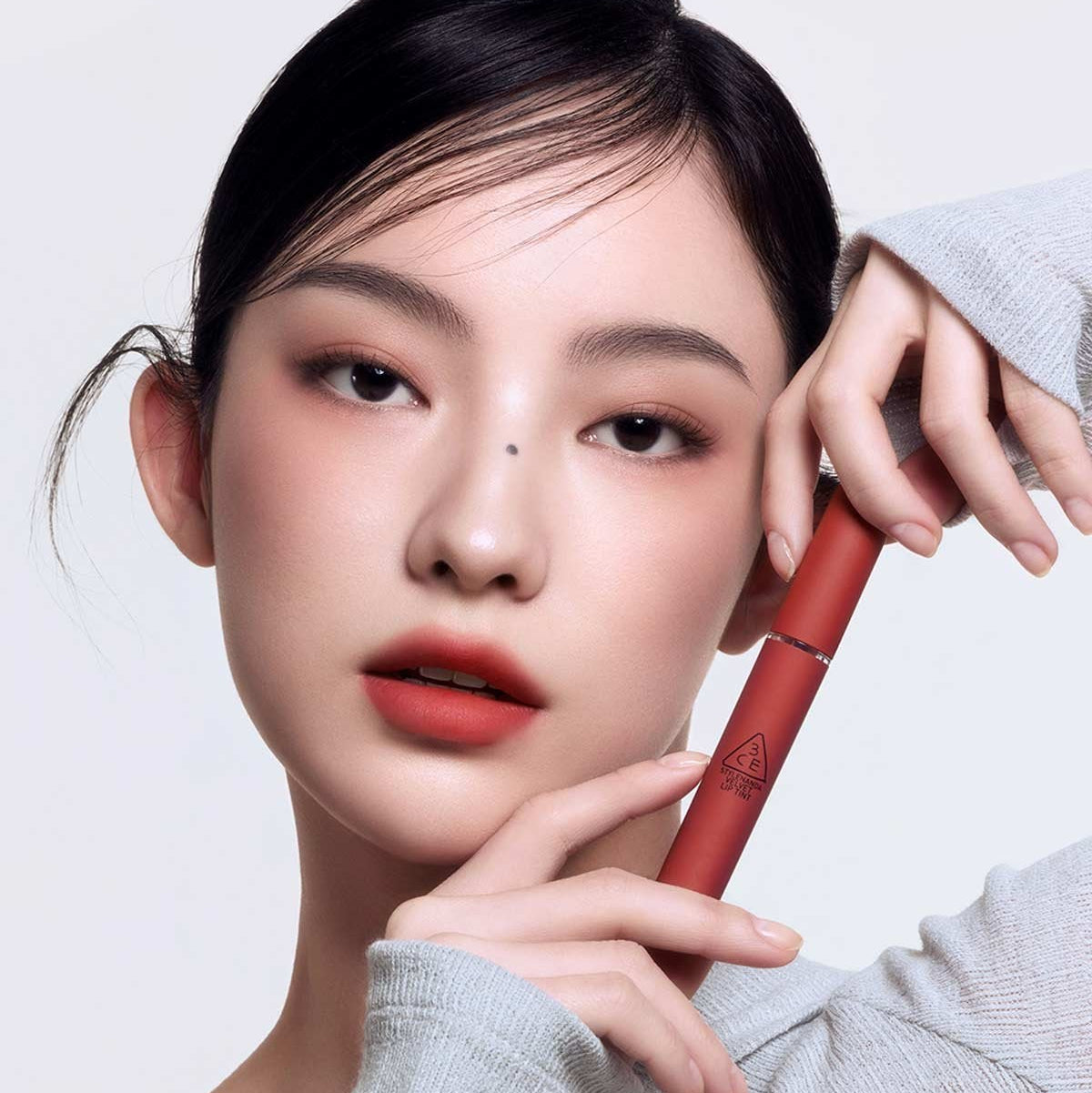 Gros plan sur 3CE Velvet Lip Tint de la marque 3CE, lèvres et tube rouge, maquillage et mains sur fond clair.