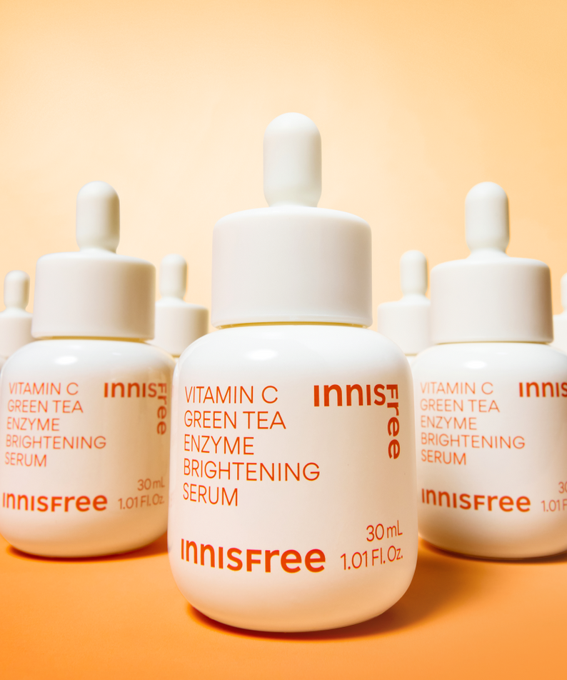 Décris l'image en français: INNISFREE Vitamin C Green Tea Enzyme Brightening Serum d’Innisfree, flacons sur fond orange.
