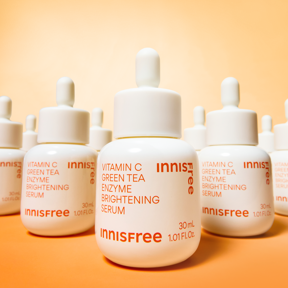 Décris l'image en français: INNISFREE Vitamin C Green Tea Enzyme Brightening Serum d’Innisfree, flacons sur fond orange.