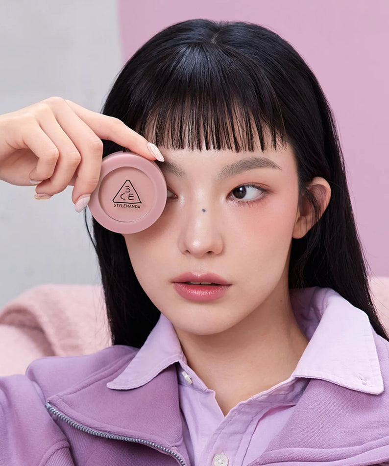 3CE Face Blush, Mood Recipe: la main applique le blush rose sur le visage d’une femme, fond pastel.