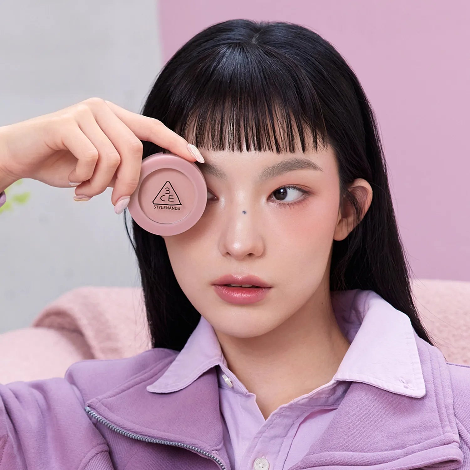 3CE Face Blush, Mood Recipe: la main applique le blush rose sur le visage d’une femme, fond pastel.