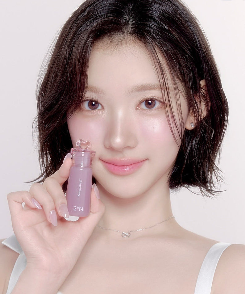 Décris l'image en français : 2AN  Heart Wearing Liquid Cheek 4.4g de la marque 2AN, la fille montre le flacon près du visage.