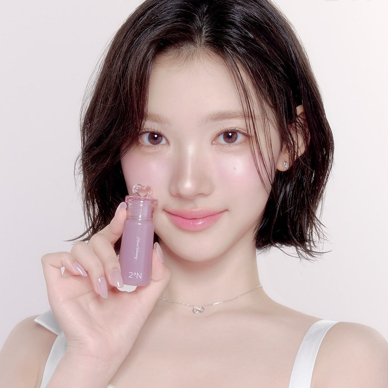 Décris l'image en français : 2AN  Heart Wearing Liquid Cheek 4.4g de la marque 2AN, la fille montre le flacon près du visage.