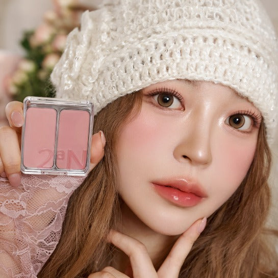 Décris l'image en français : 2AN Dual Cheek Blush de 2AN, maquillage rose sur le visage, main tenant le produit.