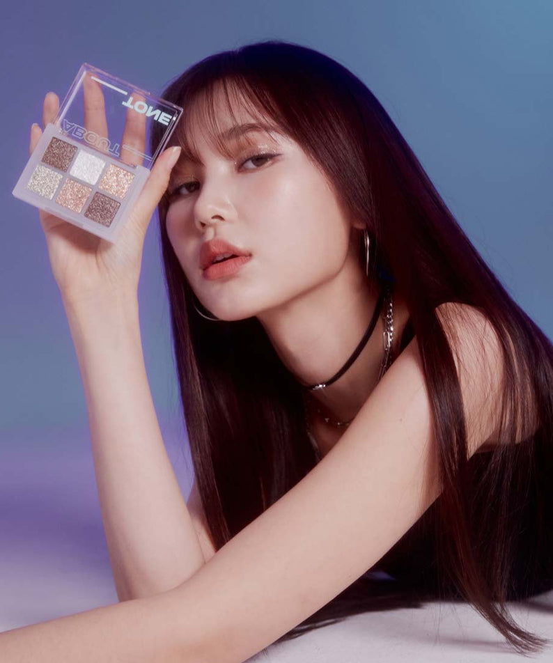 ABOUT TONE Oh My Glitter, palette glitter tenue par une femme maquillée, contexte studio bleu, maquillage lumineux.