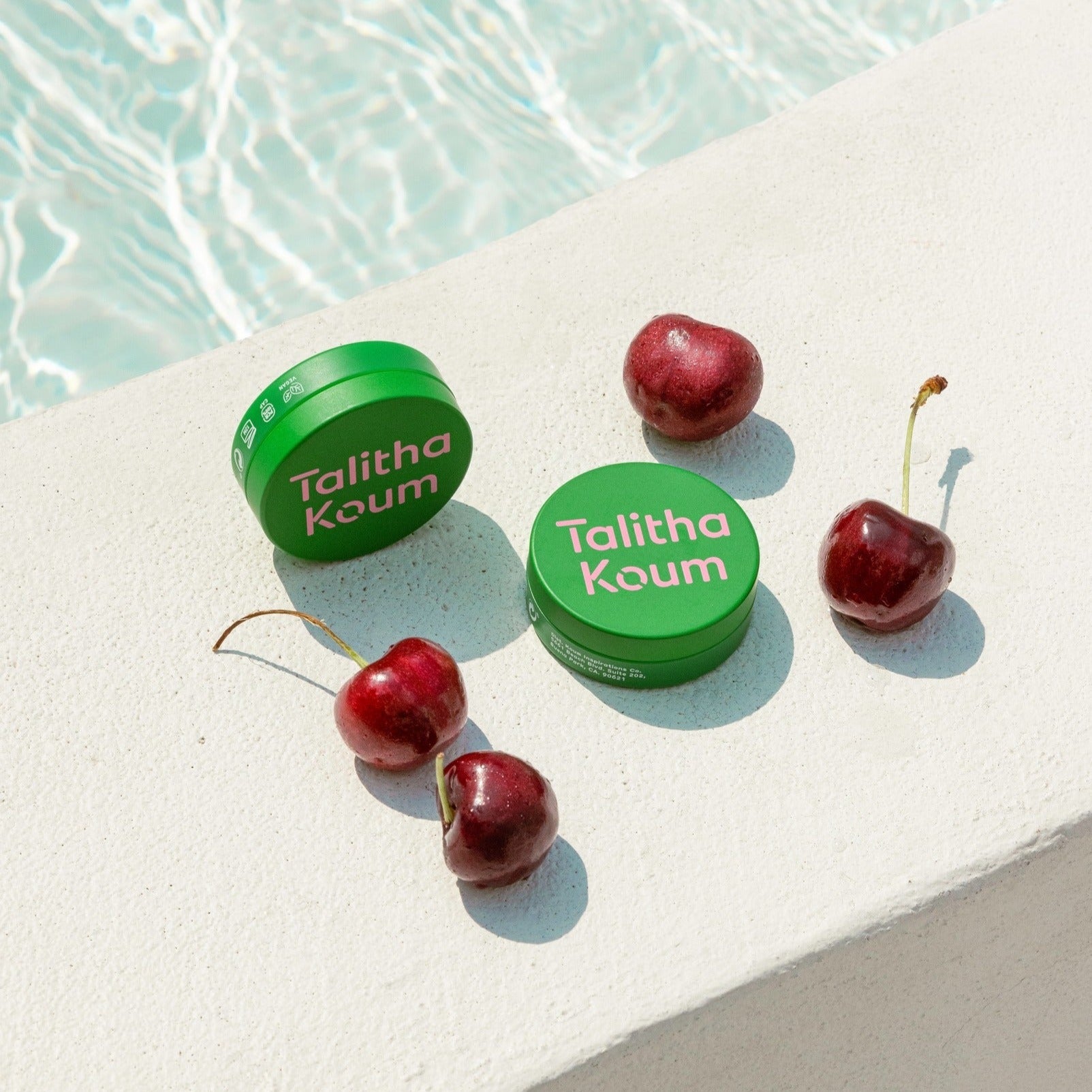 TALITHA KOUM Lipcure Balm 6g, baume à lèvres vert sur sable près de cerises, marque visible en bord de piscine.