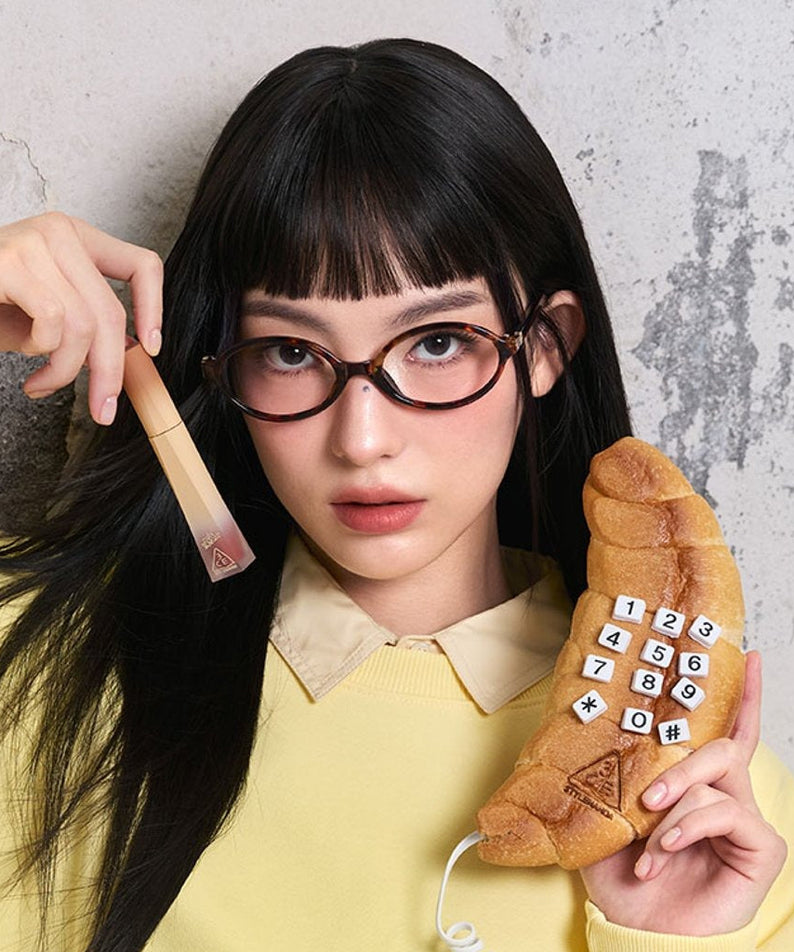 Décris l'image en français : 3CE COLOR BAKER VELVET LIP TINT PLUSH, marque 3CE, maquillage sur fond mural avec femme aux lunettes.