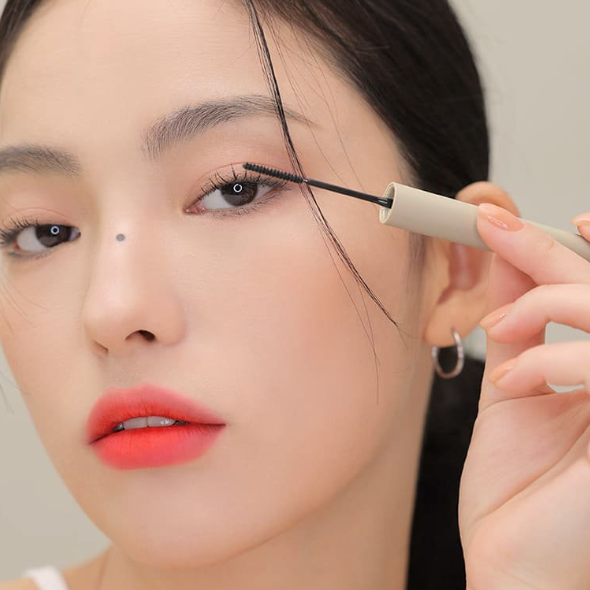 Décris l'image en français : 3CE Super Slim Waterproof Mascara, marque 3CE, maquillage des cils avec brosse fine sur fond beige.
