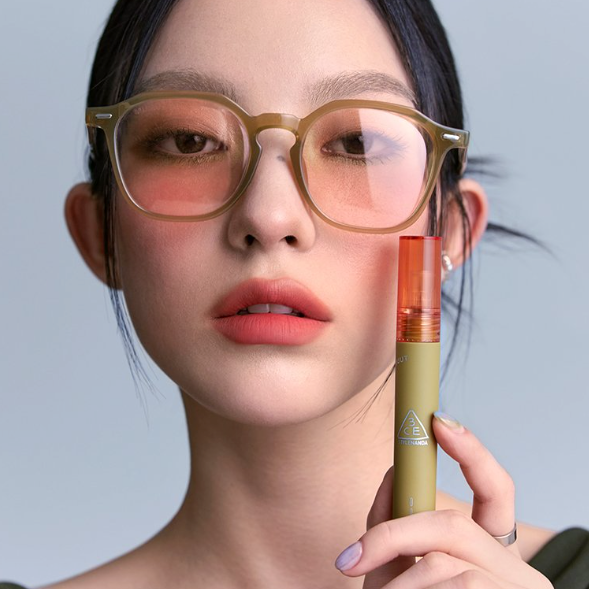 3CE Velvet Lip Tint 3 Shades, maquillage lèvres et packaging sur fond bleu, femme au regard maquillé, lunettes dorées.
