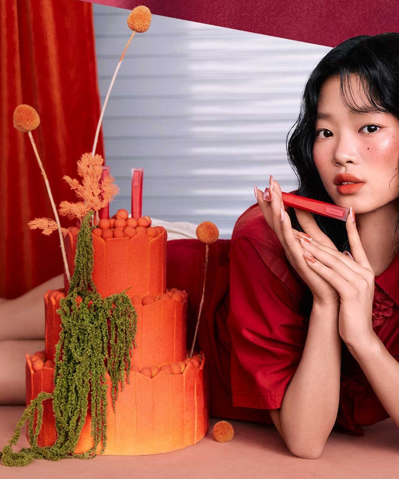 3CE Velvet Lip Tint Plush, femme maquillage lèvres rouges sur décor camaïeu rouge et orange, photo lifestyle.