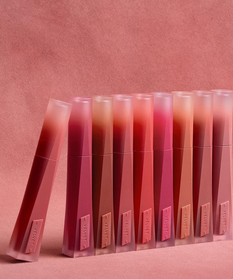 3CE Velvet Lip Tint Plush sur fond rose, rangée de tubes maquillage 3CE, rendu velouté et couleurs nude/rosées.