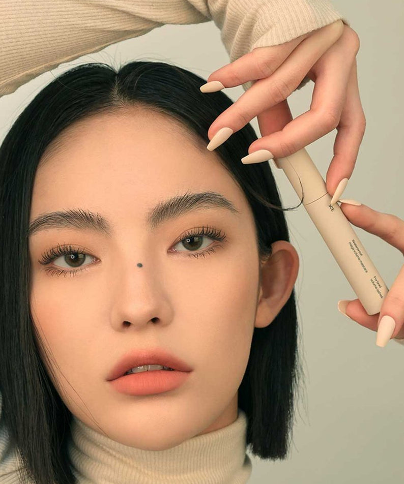 3CE Waterproof Mega Volume Mascara, flacon beige tenu près des cils d’une femme en gros plan (contexte maquillage).