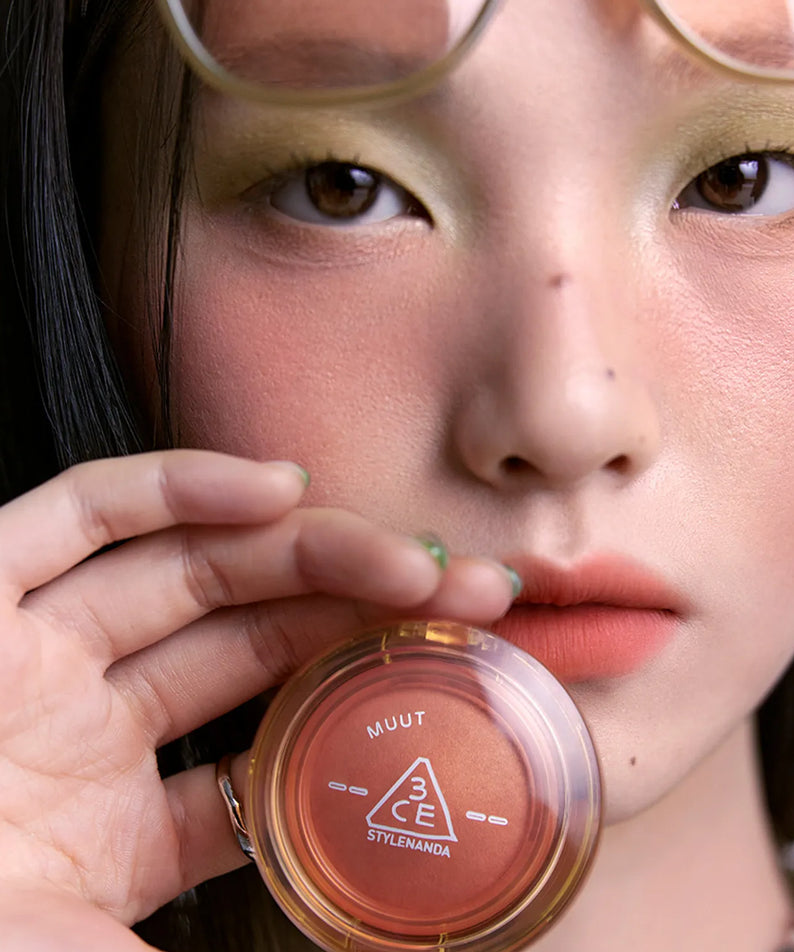 Gros plan sur le visage d'une femme tenant le 3CE Bouncy Blur Balm, maquillage estompé, peau éclatante avec le produit 3CE.