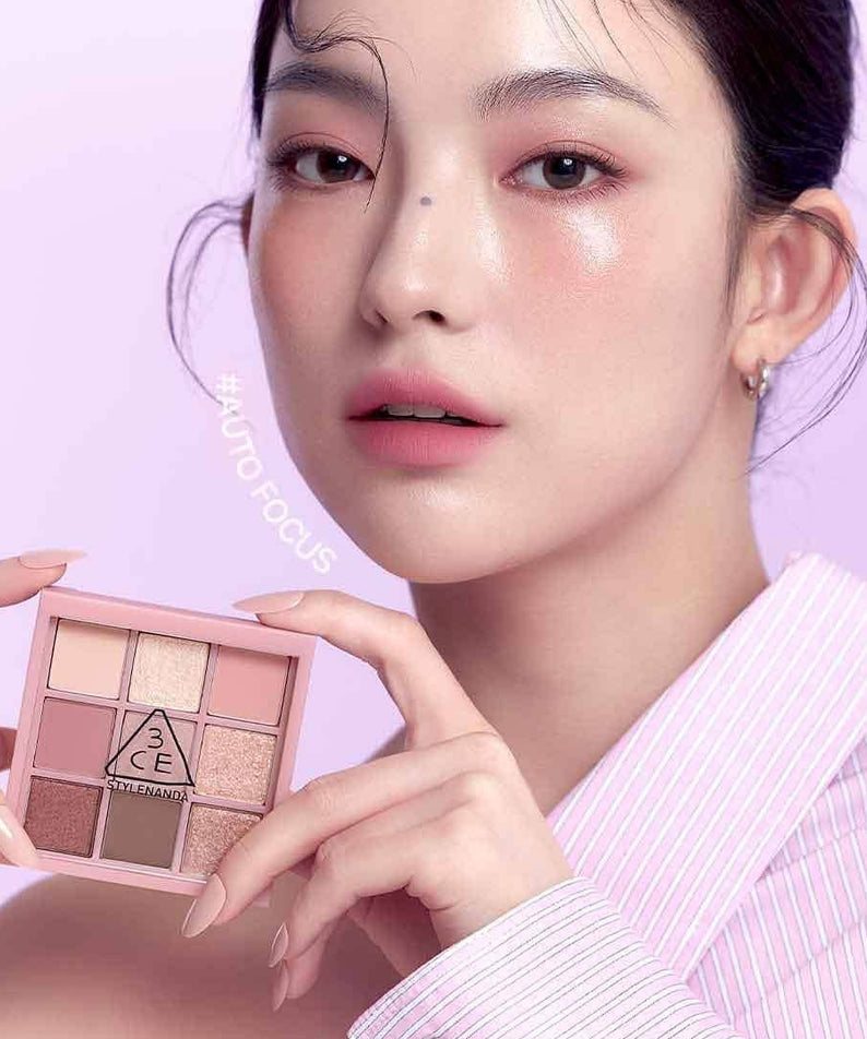 Décris l'image en français : 3CE Multi Eye Color Palette, maquillage des yeux 3CE, main tenant la palette sur fond rose clair.