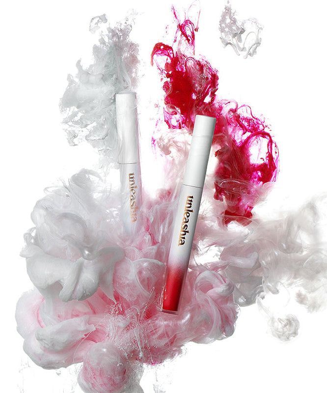 UNLEASHIA Luv Hug Velvet Tint, deux tubes maquillage roses, sur fond blanc avec nuages de fumée colorée.