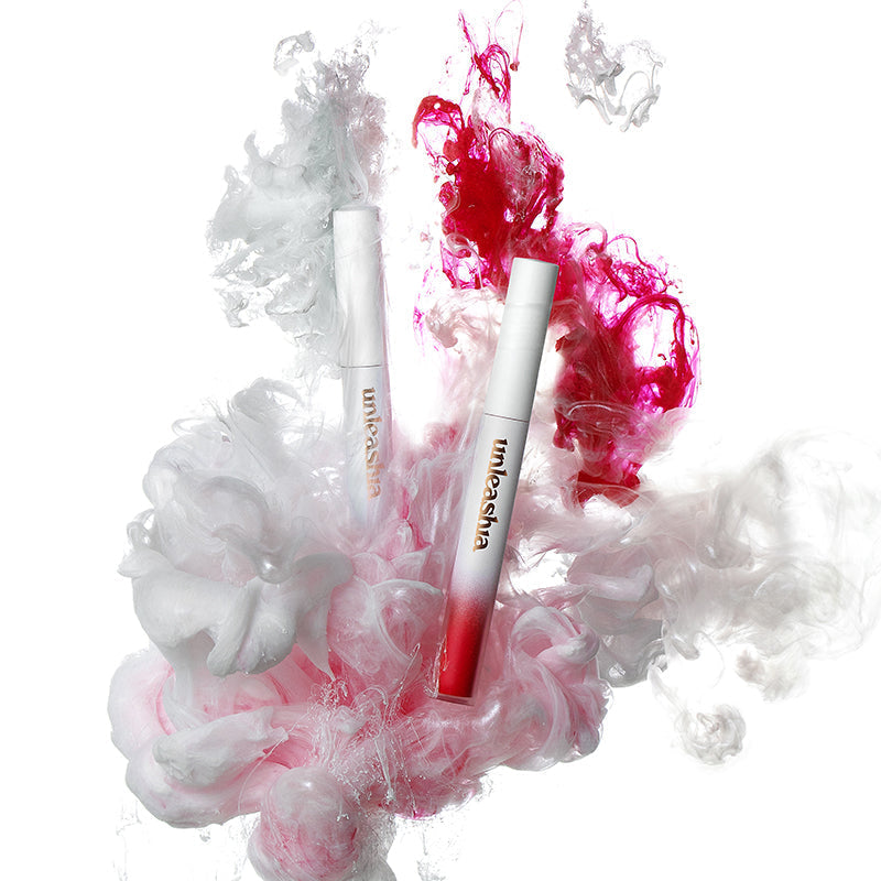 UNLEASHIA Luv Hug Velvet Tint, deux tubes maquillage roses, sur fond blanc avec nuages de fumée colorée.