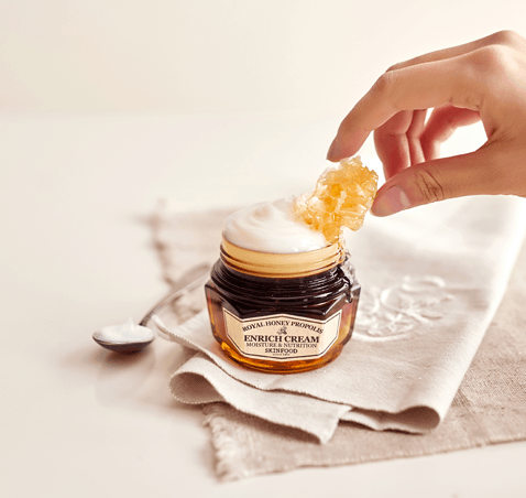 SKINFOOD Royal Honey Propolis Enrich Barrier Cream 63ml sur pot, main applique texture, ambiance soin visage sur tissu beige.
