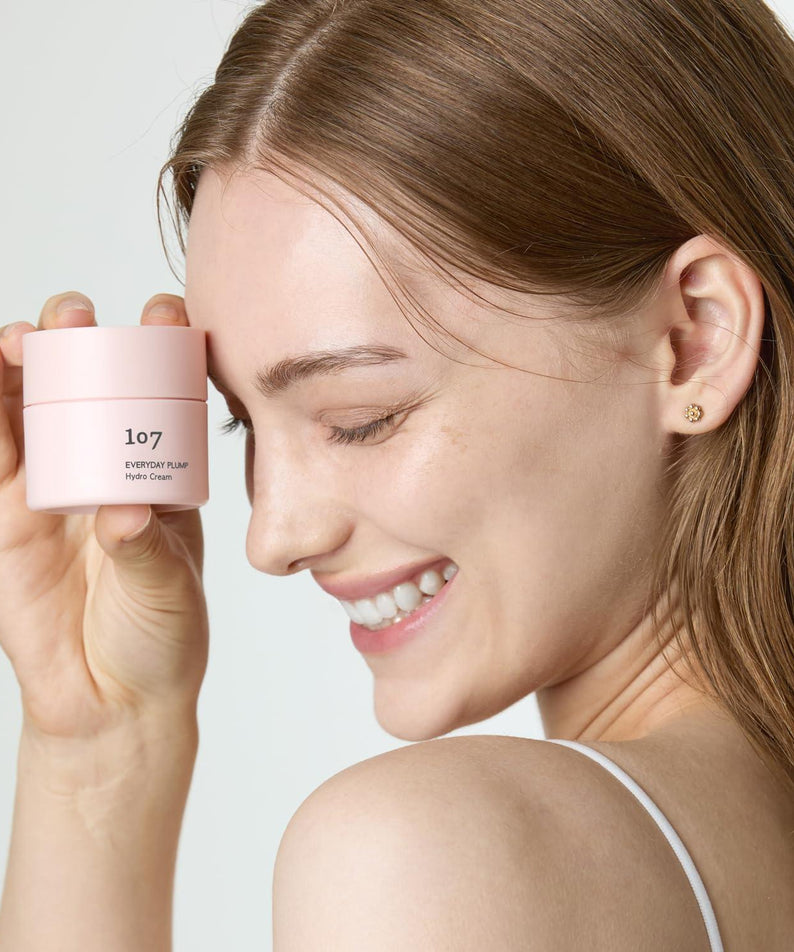 Décris l'image en français : 107 Everyday plump Hydro Cream 50ml de la marque 107, pot rose, visage souriant fond clair.