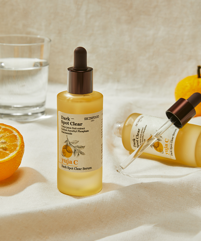 SKINFOOD Yuja C Dark Spot Clear Serum 50ml de SKINFOOD, sérum sur tissu avec oranges et flacons sur fond clair