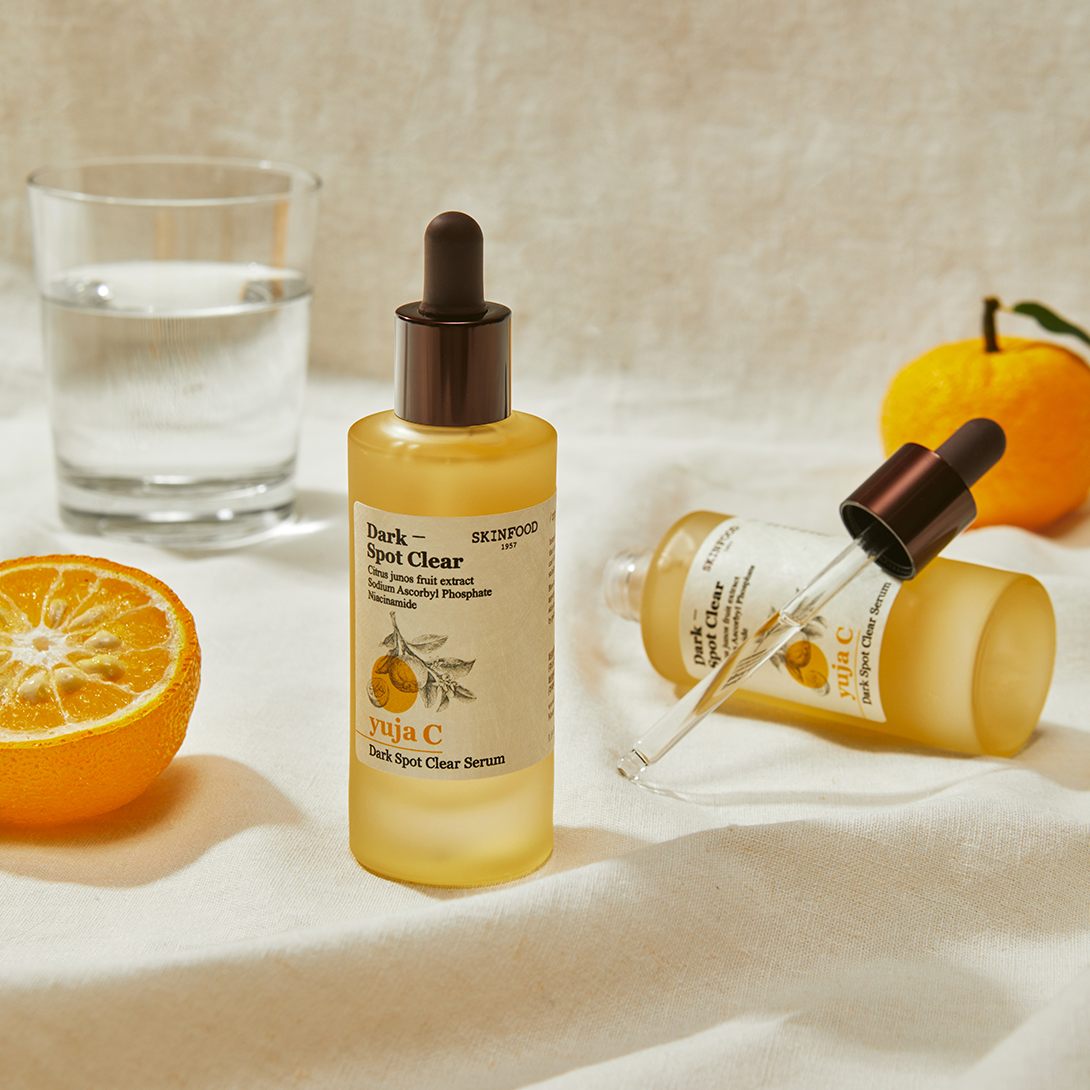 SKINFOOD Yuja C Dark Spot Clear Serum 50ml de SKINFOOD, sérum sur tissu avec oranges et flacons sur fond clair
