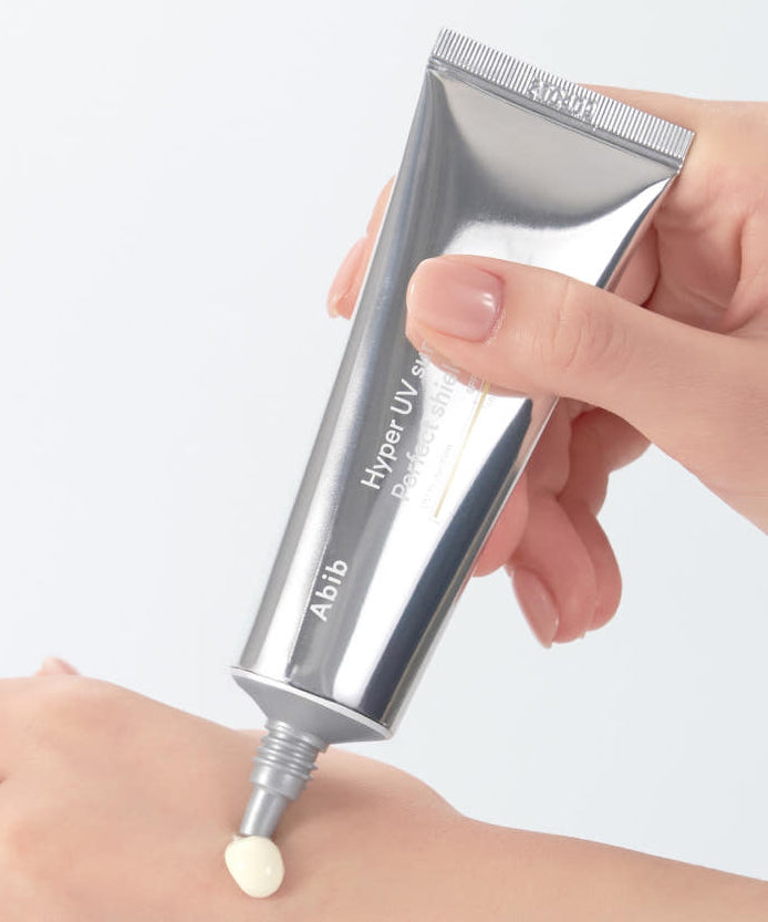 Main appliquant ABIB Hyper UV Sunscreen Perfect Shield 50ml, tube argenté sur peau, contexte solaire de protection UV.
