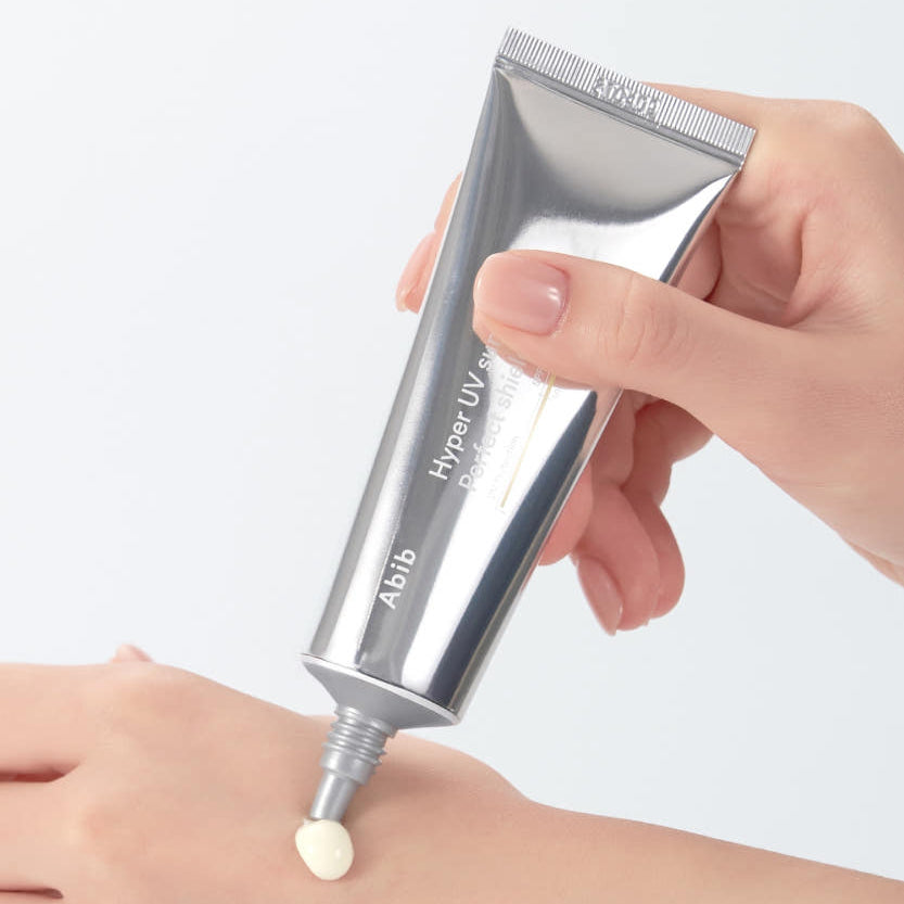 Main appliquant ABIB Hyper UV Sunscreen Perfect Shield 50ml, tube argenté sur peau, contexte solaire de protection UV.