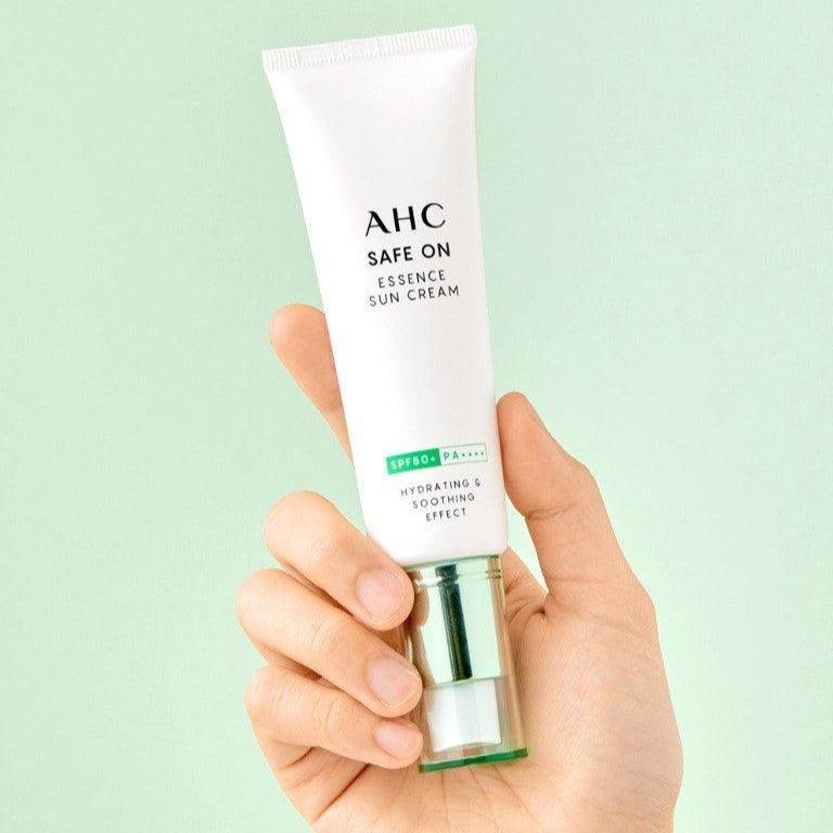 AHC Safe On Essence Sunscreen 50ml par AHC, tube tenu en main sur fond vert, contexte crème solaire.