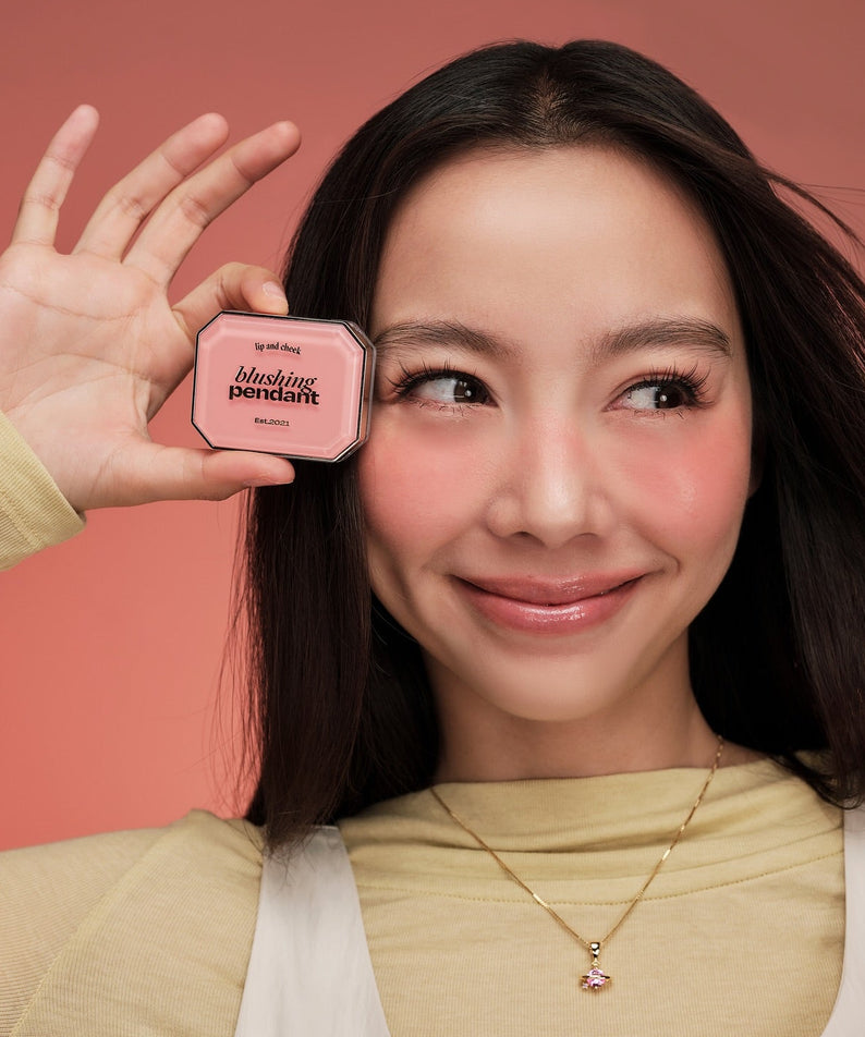 Décris l’image en français : ALTERNATIVE STEREO Blushing Pendant Lip & Cheek, femme sur fond rose tenant le produit
