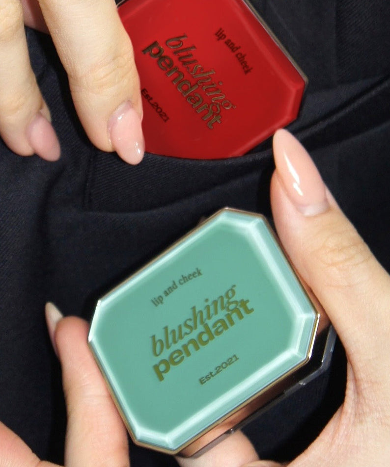 Deux poudriers ALTERNATIVE STEREO Blushing Pendant Lip & Cheeck tenus par des mains avec vernis nude, un rouge et un vert.