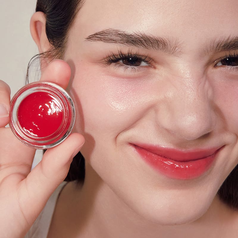 ANILLO Petal Glow Balm, baume à lèvres rouge en pot, gros plan sur visage maquillé, main tenant le produit, contexte beauté.