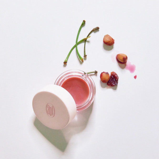 ANILLO Rose Glow Balm de la marque ANILLO, baume rose sur fond blanc avec fleurs et fruits, éclairage naturel.