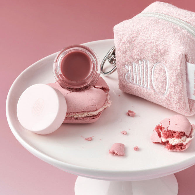 ANILLO Rose Glow Balm, baume rosé sur assiette blanche, serviette et macarons roses en fond rose, marque ANILLO.