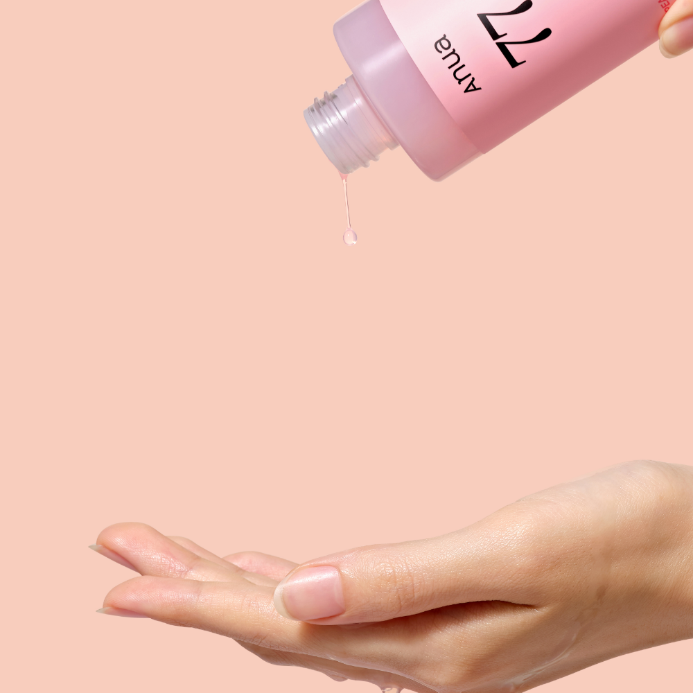 ANUA Peach 77 + Niacin Essence Toner 250ml, flacon sur peau, main applique le sérum, fond rose, marque ANUA