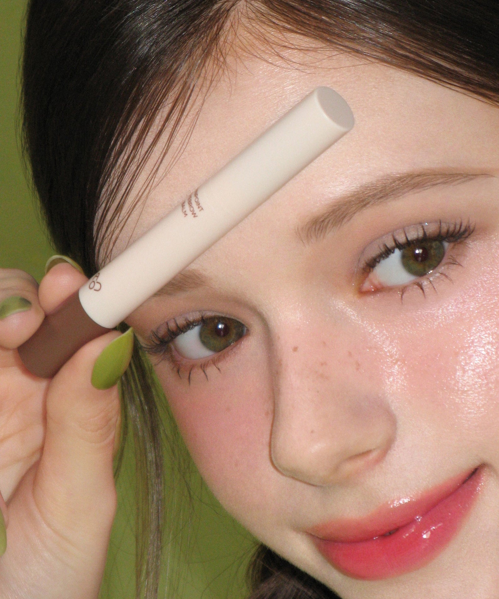 AOU Pinpoint Eyebrow Balm, tube beige sur une femme, application sur les sourcils, contexte maquillage vert.
