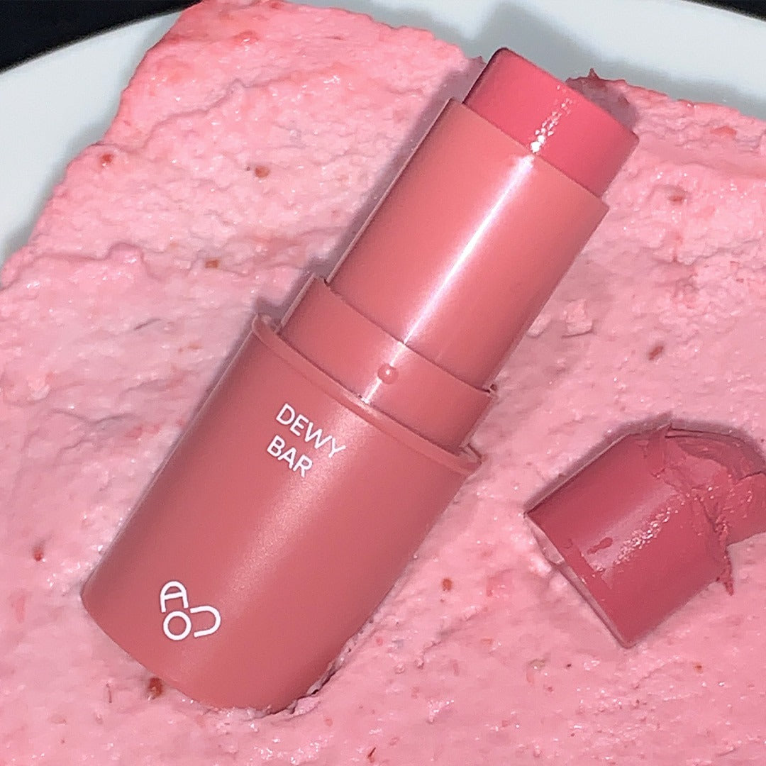 AOU Dewy Bar, marque AOU : baume hydratant rose sur fond crème texturée, capsule et étui brillants