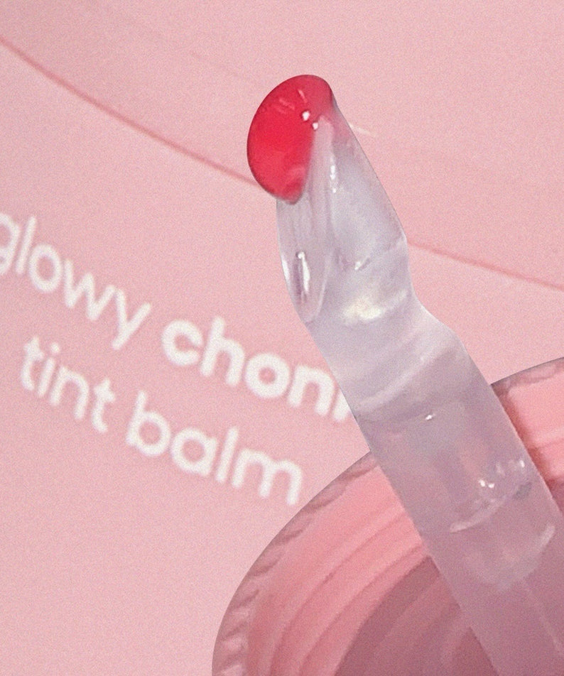 AOU Glowy Chonky Tint Balm en gros plan avec applicateur plongé dans le baume rose, fond rose pâle.